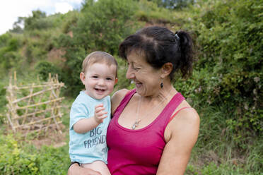 Cheerful grandmother carrying granddaughter while standing in field - GEMF04069