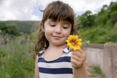 Niedliches Mädchen, das eine Blume betrachtet, während es im Freien steht - GEMF04072