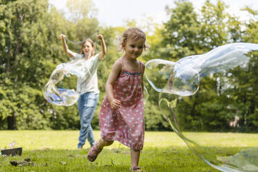 Baby girl running after bubble with mother standing in background at park - DIGF12906