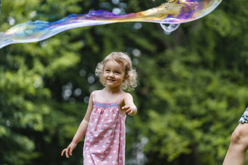 Smiling girl looking at bubble while standing at park - DIGF12911