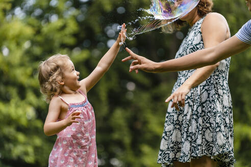 Girl exploding bubble while playing with family at park - DIGF12912