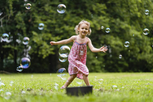Smiling girl running amidst bubbles at park - DIGF12916