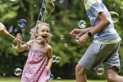 Cheerful girl enjoying with brother while running amidst bubbles at public park - DIGF12921