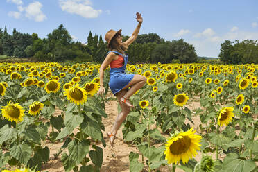 Smiling woman jumping in sunflower field during summer - VEGF02712