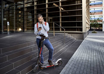 Businessman texting through phone while standing with electric scooter against staircase - VEGF02722