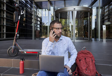 Businessman with laptop talking on phone while sitting on staircase - VEGF02730