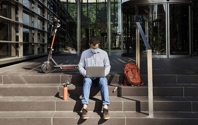 Entrepreneur with face mask using laptop on staircase during pandemic - VEGF02740