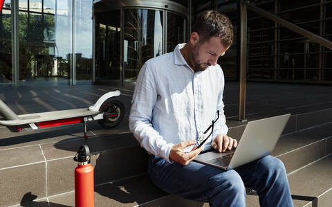 Geschäftsmann mit Brille, der über einem Laptop arbeitet, während er auf einer Treppe sitzt, lizenzfreies Stockfoto