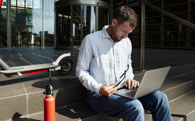 Geschäftsmann mit Brille, der über einem Laptop arbeitet, während er auf einer Treppe sitzt - VEGF02741