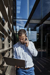 Entrepreneur talking on phone while holding laptop against building in city - VEGF02747