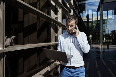 Entrepreneur talking on phone while holding laptop in city - VEGF02748