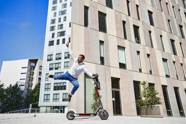Geschäftsmann springt, während er einen Motorroller in der Stadt an einem sonnigen Tag hält - VEGF02755
