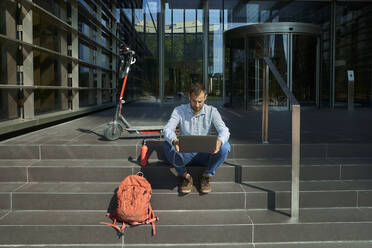 Entrepreneur with in-ear headphones using laptop while sitting on staircase - VEGF02761