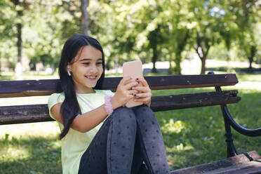 Lächelndes Mädchen, das auf einer Bank im Park sitzt und telefoniert - MOMF00891