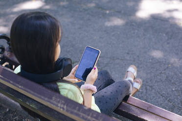 Mädchen mit Kopfhörer, das auf einer Bank im Park sitzt und die Technik nutzt - MOMF00894