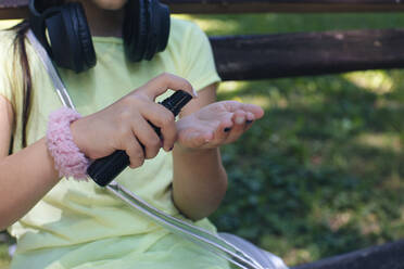 Girl using hand sanitizer while sitting on bench in park - MOMF00898