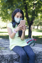 Mädchen mit Gesichtsmaske telefoniert in einem öffentlichen Park - MOMF00899
