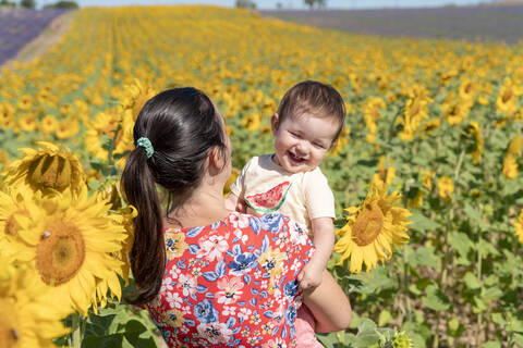 Mutter trägt fröhliche Tochter in einem Sonnenblumenfeld im Sommer, lizenzfreies Stockfoto