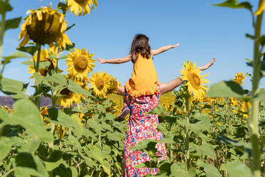 Mutter trägt Tochter auf den Schultern in einem Sonnenblumenfeld gegen den klaren Himmel - GEMF04087