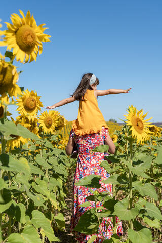 Mutter trägt Tochter auf den Schultern in einem Sonnenblumenfeld vor blauem Himmel, lizenzfreies Stockfoto