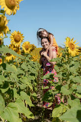 Smiling mother carrying daughter on shoulders in sunflower field - GEMF04089