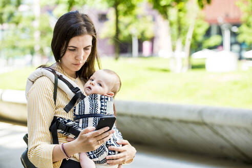 Mutter mit ihrem kleinen Jungen in der Babytrage, der sein Smartphone in der Stadt benutzt - IHF00397