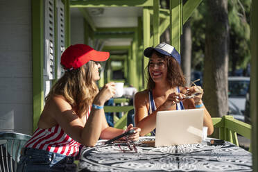 Glückliche Frauen genießen das Wochenende, während sie mit Kaffee und Laptop in einem Café an einem sonnigen Tag sitzen - OCMF01611