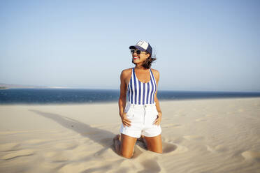 Smiling woman kneeling on sand while looking away at beach against clear blue sky - OCMF01619