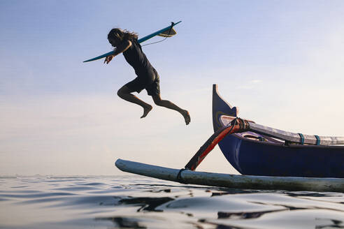 Mann mit Surfbrett springt in Meer gegen klaren blauen Himmel - KNTF05187