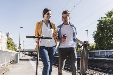 Lächelnde Frau, die einem männlichen Freund beim Spaziergang am Bahnhof ihr Smartphone zeigt - UUF20861