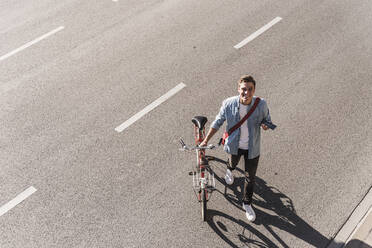 Lächelnder Mann mit Fahrrad beim Überqueren der Straße in der Stadt an einem sonnigen Tag - UUF20867