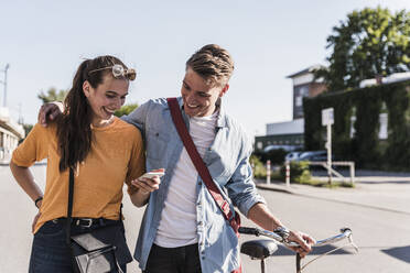 Happy young woman showing smartphone to boyfriend while walking in city - UUF20870