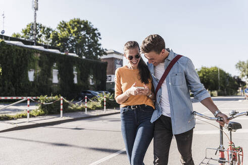 Fröhliches junges Paar schaut auf sein Smartphone, während es auf der Straße in der Stadt spazieren geht - UUF20871