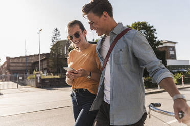 Smiling young couple looking at mobile phone while walking on street in city - UUF20873