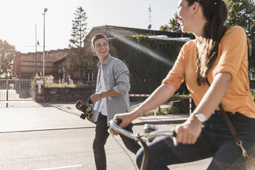 Cheerful young man looking at girlfriend riding bicycle on street in city during sunny day - UUF20874