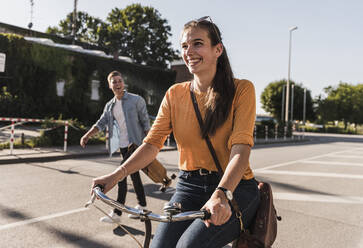 Glückliche junge Frau fährt Fahrrad, während ihr Freund auf der Straße in der Stadt spazieren geht - UUF20875