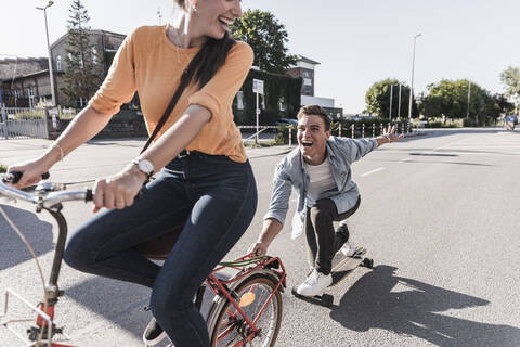 Fröhliche junge Frau fährt Fahrrad, während ihr Freund auf einer Straße in der Stadt Skateboard fährt, lizenzfreies Stockfoto