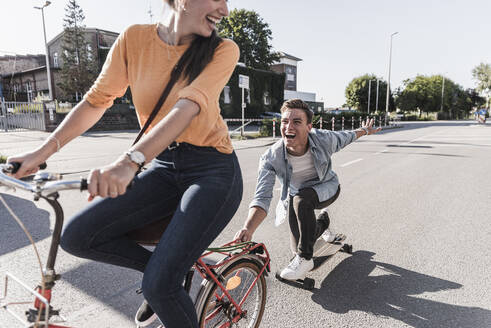 Fröhliche junge Frau fährt Fahrrad, während ihr Freund auf einer Straße in der Stadt Skateboard fährt - UUF20876