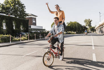 Fröhliche junge Frau steht hinter ihrem Freund, der auf der Straße Fahrrad fährt - UUF20878