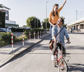 Glückliche Frau steht mit erhobener Hand hinter ihrem Freund, der auf einer Straße in der Stadt Fahrrad fährt - UUF20879