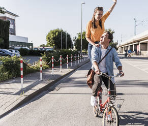 Happy woman standing with hand raised behind boyfriend riding bicycle on street in city - UUF20879