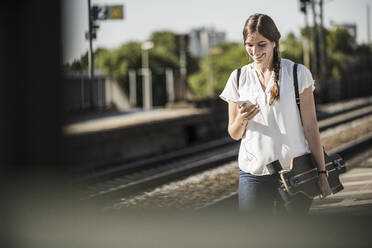 Lächelnde junge Frau mit Skateboard und Smartphone am Bahnhof - UUF20888