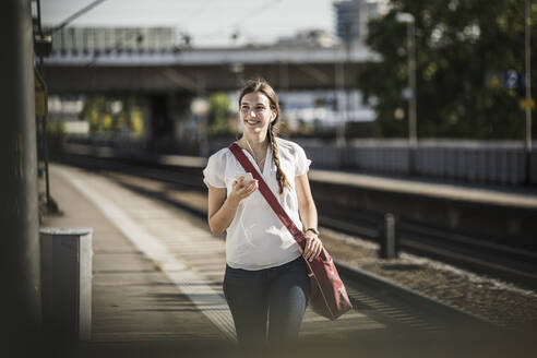 Lächelnde Frau hört Musik über ihr Mobiltelefon, während sie auf dem Bahnhof spazieren geht - UUF20893