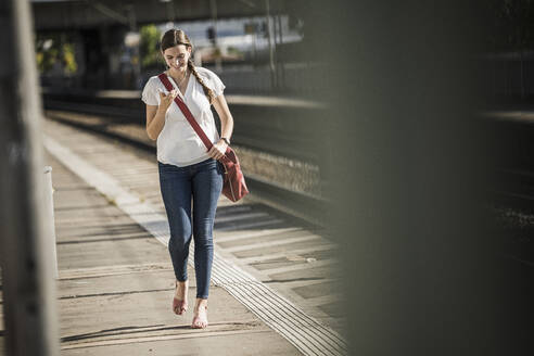 Junge Frau hört Musik über ihr Handy, während sie auf dem Bahnsteig spazieren geht - UUF20896