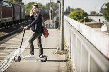 Young man talking on mobile phone while standing with push scooter at railroad station platform - UUF20897