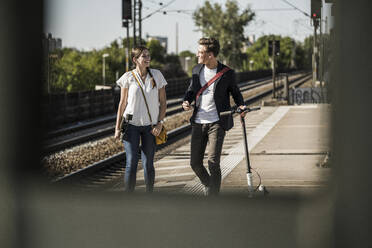 Smiling male and female friends talking while walking on railroad station platform - UUF20903