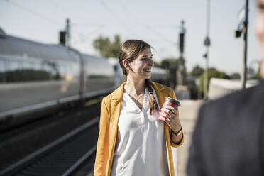 Junge Frau mit wiederverwendbarem Kaffeebecher, die am Bahnhof steht und wegschaut - UUF20907