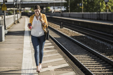 Lächelnde junge Frau, die ein Smartphone benutzt, während sie auf dem Bahnsteig spazieren geht - UUF20910
