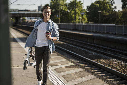 Lächelnder junger Mann, der einen Roller trägt, während er auf dem Bahnsteig spazieren geht - UUF20914