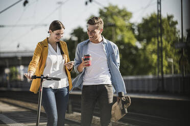 Junge Frau zeigt einem männlichen Freund beim Spaziergang am Bahnhof ihr Smartphone - UUF20915